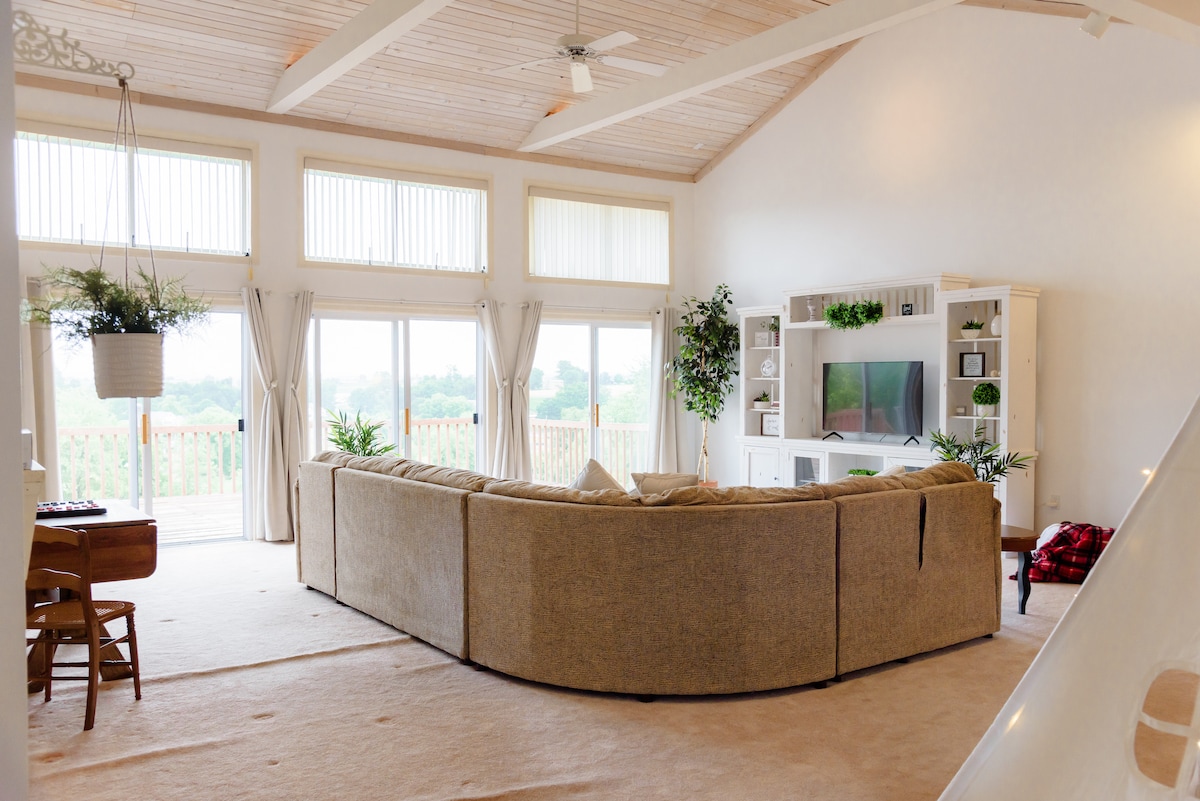 A spacious living area features a large, curved sofa facing a media center, surrounded by green plants. High cathedral ceilings and ample natural light filter through oversized windows, with views of the balcony and scenic farmland visible in the background.