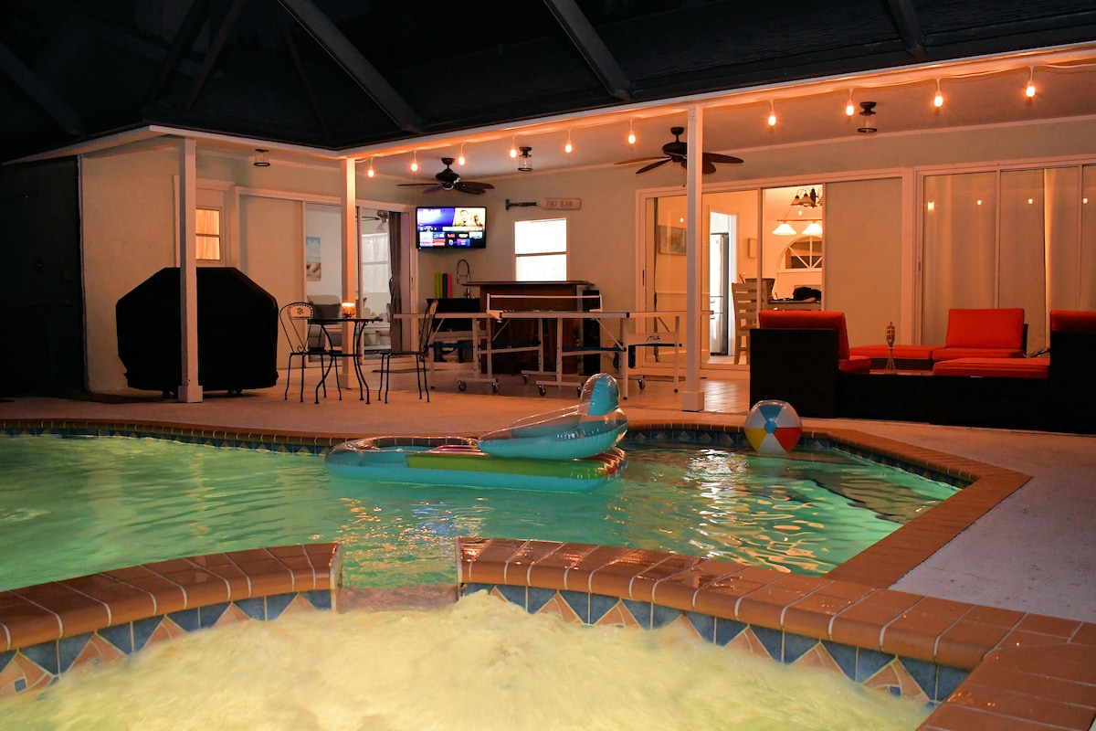 A sparkling pool is visible, accompanied by a hot tub featuring a waterfall. Colorful pool floats lounge on the water's surface. The outdoor space connects to an enclosed patio area, showcasing seating and a nearby TV, with additional features detectable through expansive sliding glass doors.
