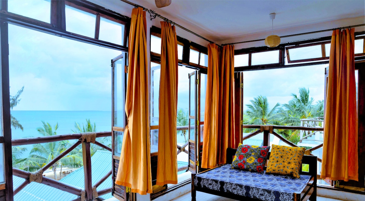 A well-lit indoor space features large windows adorned with bright yellow curtains, providing panoramic views of the ocean and palms. A cozy seating area is highlighted by a patterned cushion arrangement on a low bench, inviting relaxation and enjoyment of the scenic surroundings.