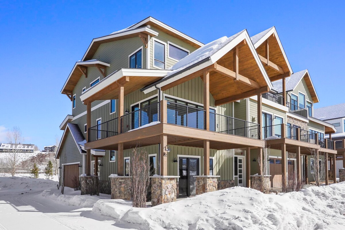 A multi-level home is shown from the outside, featuring a large deck with wooden railings and a stone base. Snow covers the ground, and large windows are visible, providing natural light. The roof is angled with overhangs, adding dimension to the structure.