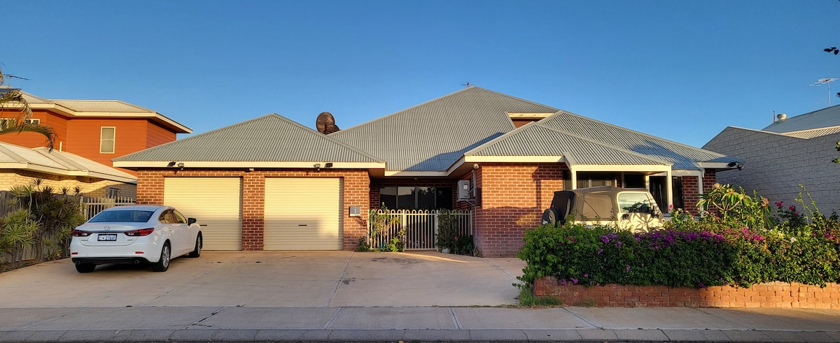 The exterior of a spacious, single-story home is presented, featuring warm brick walls and a light gray roof. A neatly paved driveway accommodates two parked vehicles, and vibrant greenery is positioned along the front, adding a touch of color to the setting.