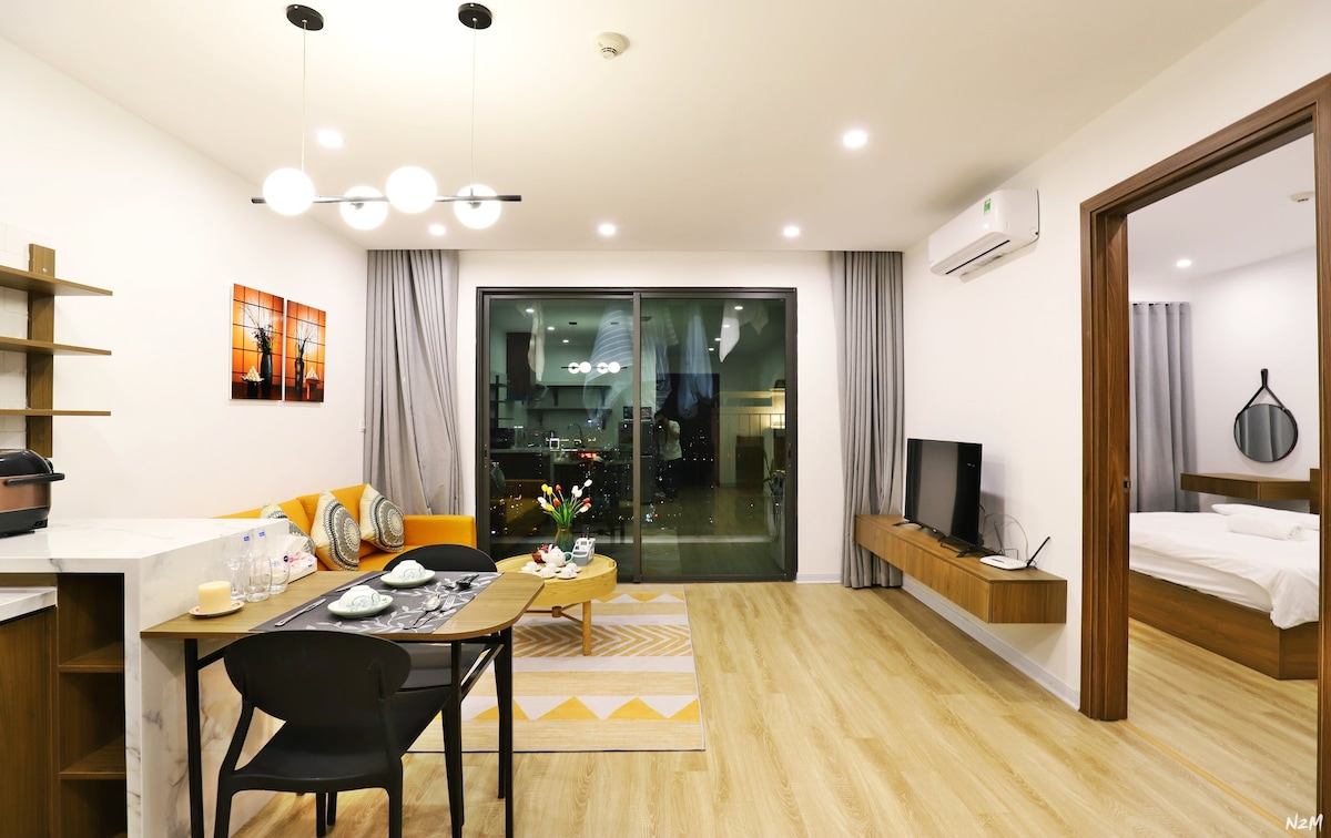 A modern living area showcases a comfortable orange sofa paired with a sleek dining table set for four. Large glass doors provide access to a balcony, while minimalist décor and soft lighting enhance the space. A mounted television and warm wood accents create a cozy environment.