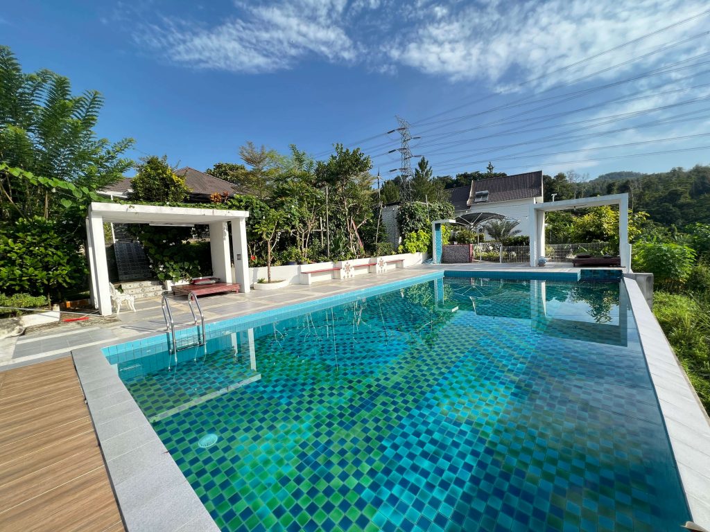 An infinity pool is surrounded by lush greenery, offering a serene environment. Light blue and green mosaic tiles create a vibrant pattern on the water's surface. White lounge chairs are positioned nearby, along with shaded areas and comfortable seating, providing an inviting space for relaxation.