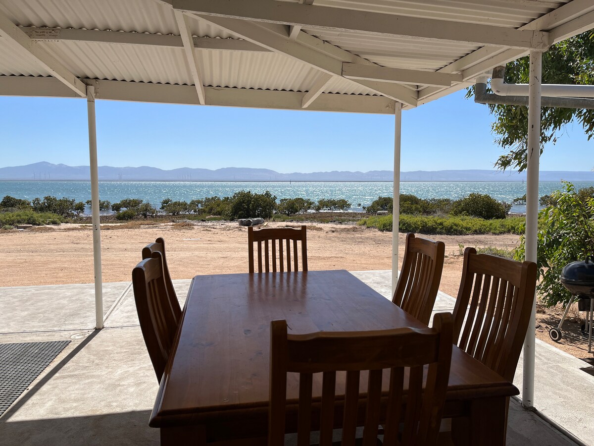 A dining area is positioned under a shaded outdoor patio, featuring a wooden table surrounded by six chairs. The view overlooks Spencer Gulf, showcasing water shimmering in sunlight and the distant outline of hills along the horizon.