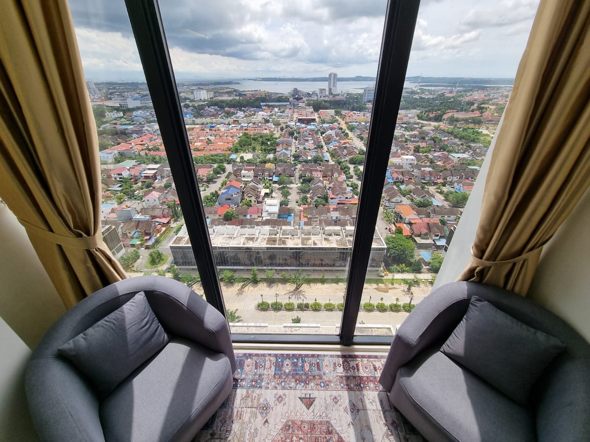 Two comfortable chairs are positioned beside a large floor-to-ceiling window, offering a panoramic view of the surrounding neighborhood and the sea in the distance. Soft curtains frame the window, with a decorative rug placed on the floor beneath.