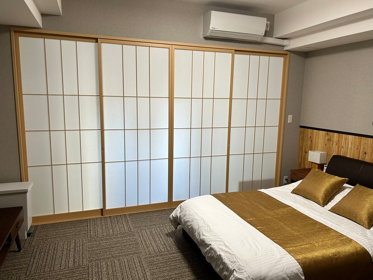 A well-appointed room showcases a spacious bed dressed in crisp linens with gold accents. A multi-panel shoji screen adds traditional charm, while an air conditioning unit is mounted above for comfort. Soft lighting and neutral wall tones create a serene atmosphere.