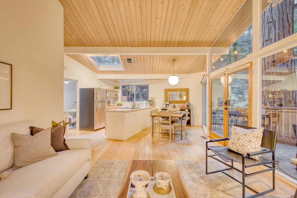 An open-concept living space features wooden ceilings, allowing natural light to flow in through skylights. The dining area is seen with a table for six, and the kitchen includes stainless steel appliances. Soft seating is arranged around a coffee table, creating a comfortable gathering spot.