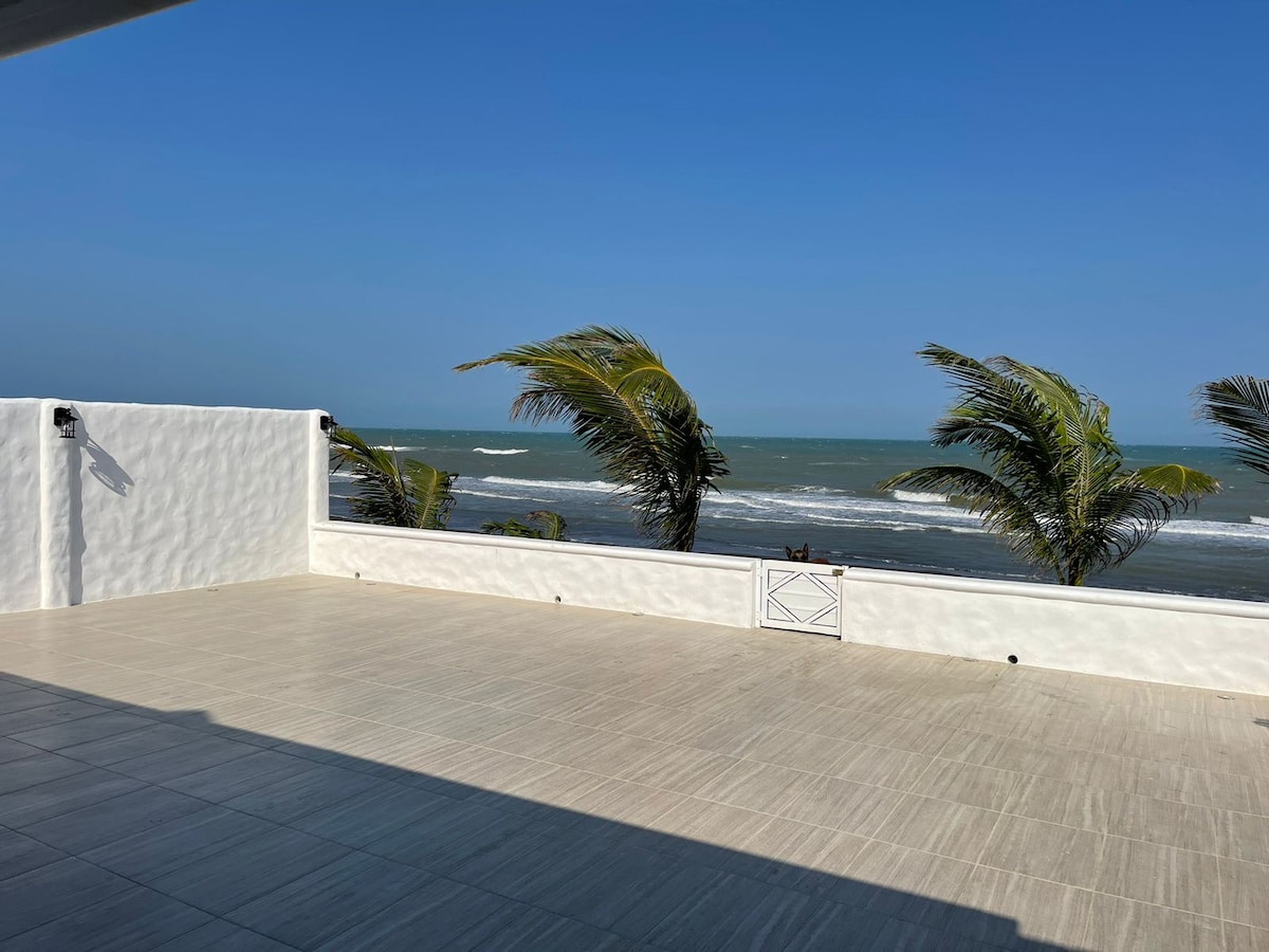 An open terrace extends toward the ocean, featuring a smooth tiled surface. Palm trees sway gently in the breeze, while waves break at the shoreline in the distance under a clear blue sky.