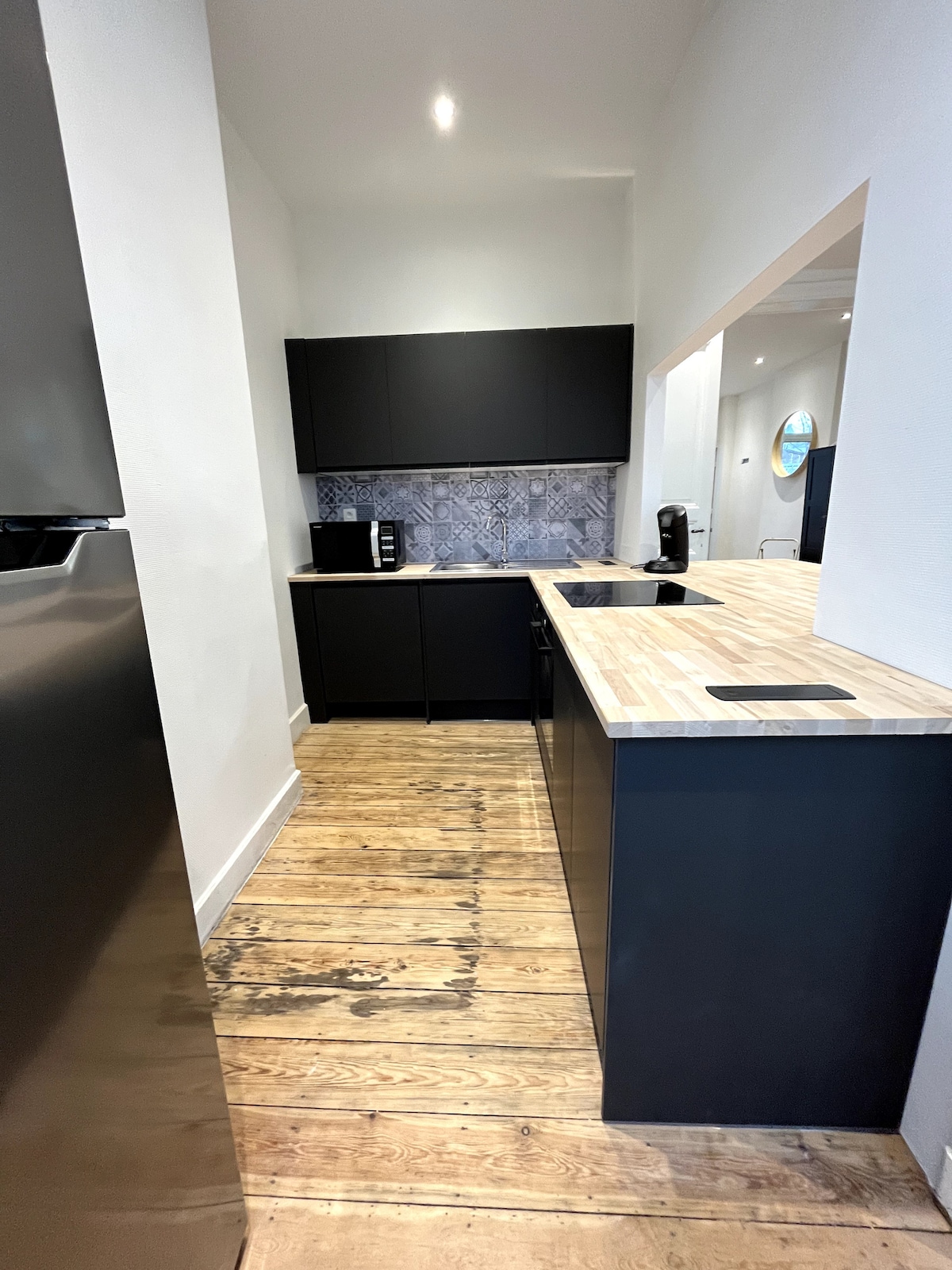 A sleek kitchen is presented with dark cabinetry and a light wood countertop. A patterned backsplash adds visual interest. The space features modern appliances, with an open layout allowing for easy movement. Natural light illuminates the wood floor, enhancing the clean aesthetic.