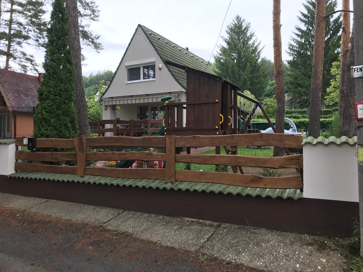 A two-story house is framed by a wooden fence, providing a welcoming entrance. Lush trees and greenery surround the property, while a playset is visible in the yard, indicating a family-friendly environment. The sloped roof adds character to the structure.