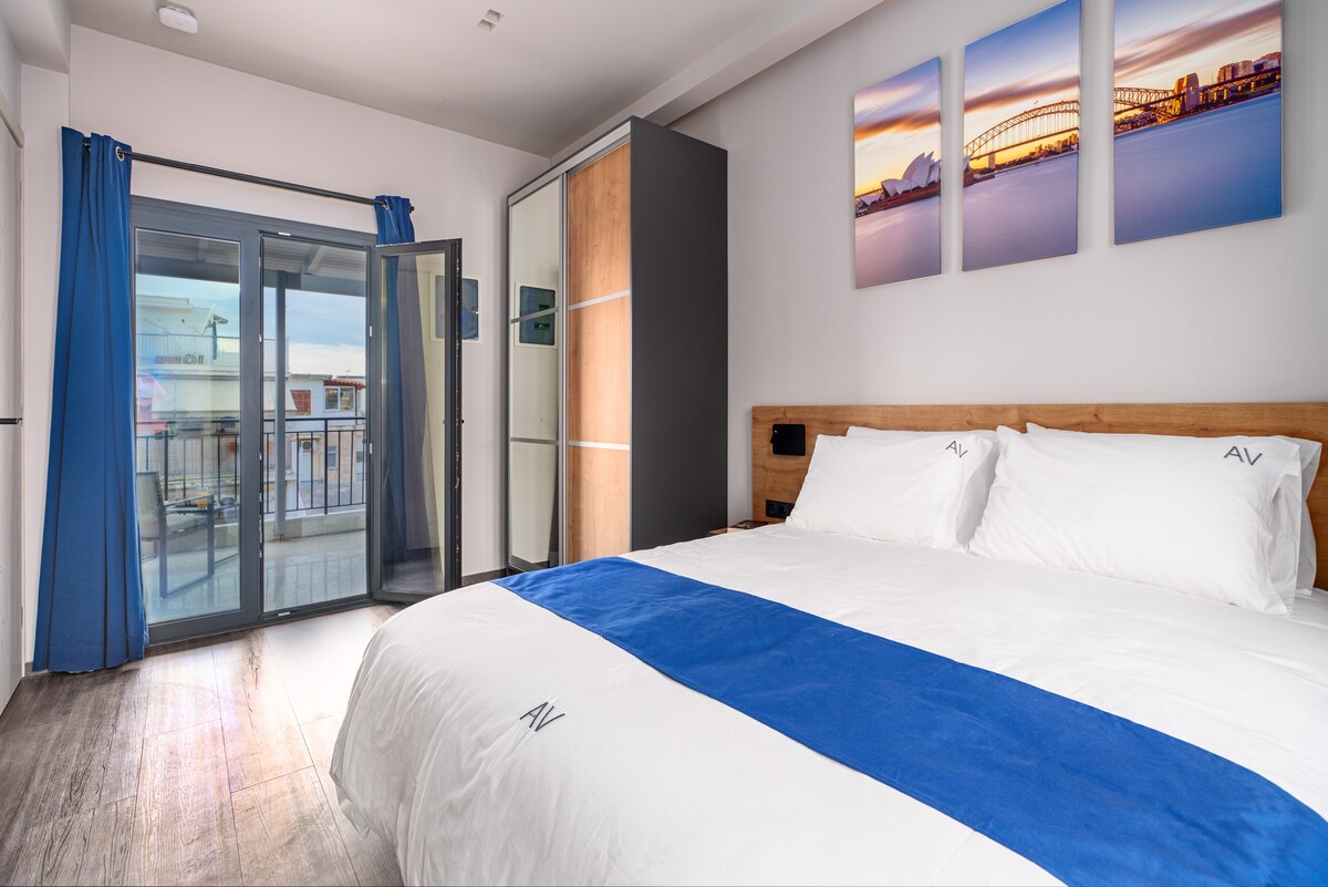 A spacious room features a large bed with crisp white linens and a blue accent blanket. Floor-to-ceiling windows allow natural light to fill the space, with sliding doors leading to a balcony. Artwork depicting a city skyline adorns the wall above the bed.