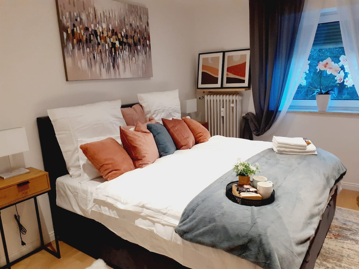 A spacious bedroom features a large double bed adorned with a mix of decorative pillows in warm tones. Two bedside lamps sit on either side of a wooden nightstand. Framed artwork and a potted orchid add subtle ambiance, while a soft throw blanket rests at the foot of the bed.