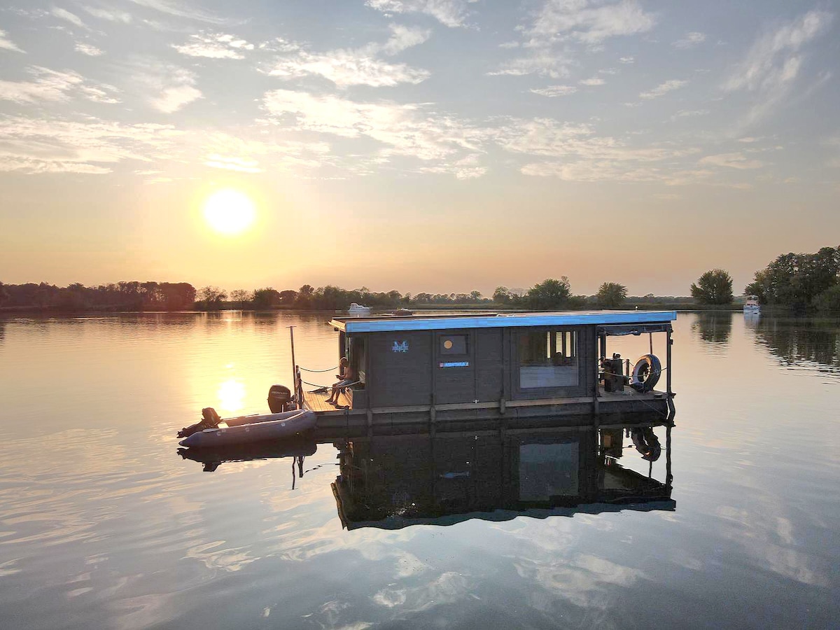A water bungalow floats serenely on the calm lake, reflecting the warm hues of a setting sun. The structure features a covered porch and is surrounded by nature, creating a peaceful and inviting atmosphere for guests.