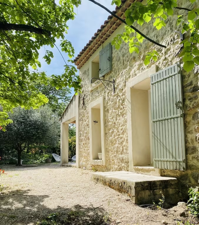 A stone house featuring rustic architecture is surrounded by greenery. Window shutters in soft green add character to the exterior, while a covered area provides a transition to the outdoor space. A path leads to a serene garden setting with seating options nearby.