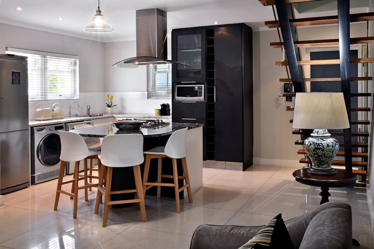 A modern kitchen and dining area feature a central island with white seating and a sleek black cabinetry. Stainless steel appliances are complemented by natural light streaming through windowed blinds, while decorative elements like a lamp add a personal touch.