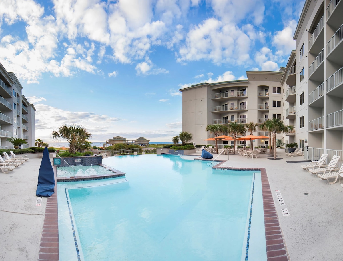 A spacious outdoor pool area features clear blue water surrounded by lounge chairs and palm trees. The nearby building offers balconies overlooking the pool. A vibrant sky is visible, with a mix of clouds and sunlight enhancing the resort's inviting atmosphere.