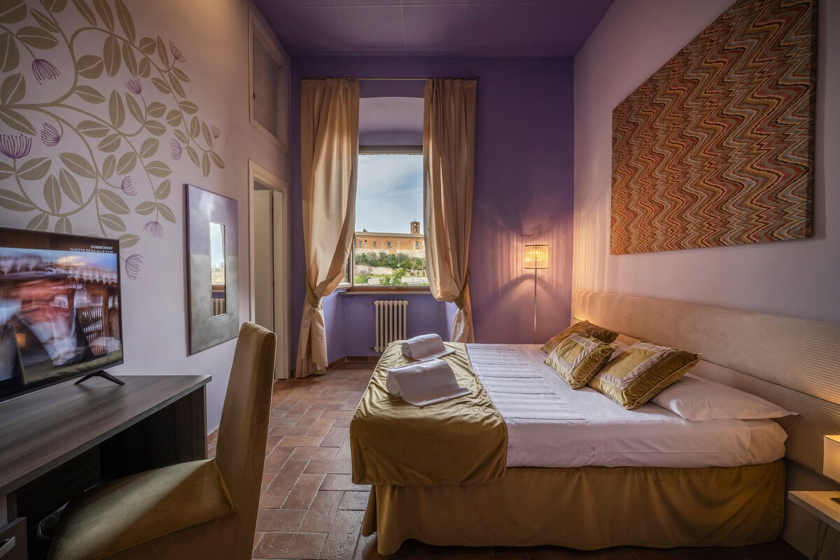 A bright and spacious guest room is presented with a large window dressed in soft curtains, providing natural light. A comfortable bed with decorative pillows is featured, accompanied by a flat-screen TV and a desk. Walls are decorated with subtle patterns, enhancing a serene ambiance.