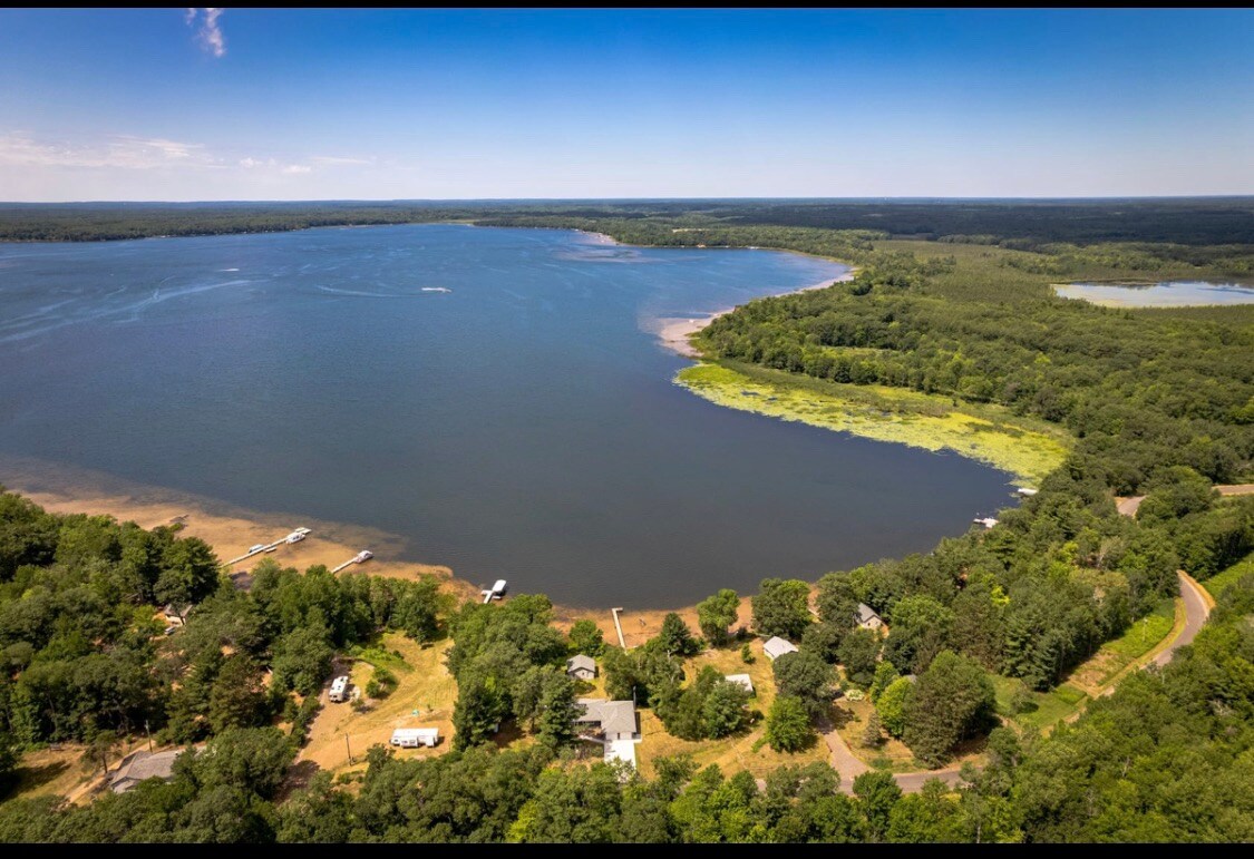 An aerial view captures a serene lake bordered by lush greenery. The coastline features a blend of sandy and rocky areas, with boats visible in the water. The expansive landscape reflects a peaceful natural setting, inviting outdoor activities.