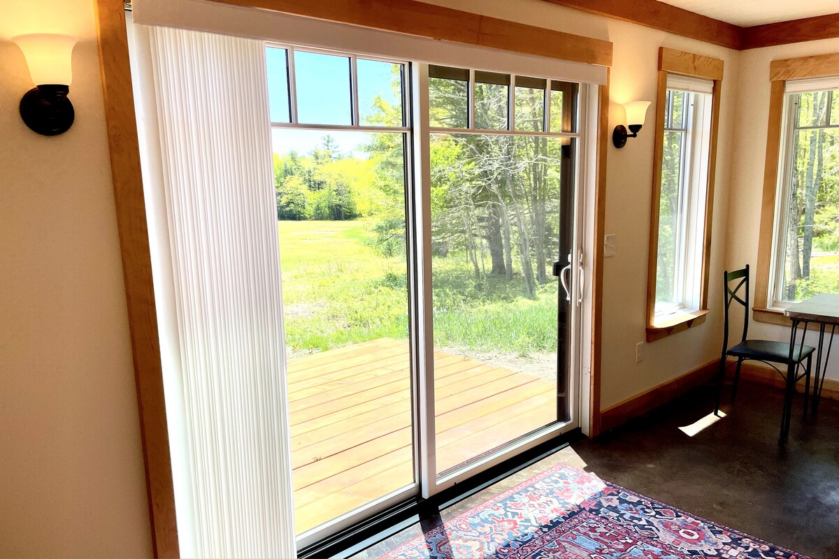 Natural light floods the room through an 8 ft sliding door, which opens to a wooden deck surrounded by greenery. The concrete-stained floor is complemented by a colorful area rug. Nearby, a small dining table is visible next to a chair, enhancing the functional space.