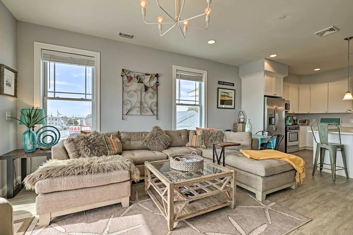 A bright living area features a spacious sectional sofa adorned with colorful cushions, complemented by a patterned area rug. Large windows provide natural light, while a stylish chandelier hangs above a coffee table. A kitchen area with modern appliances is visible in the background.