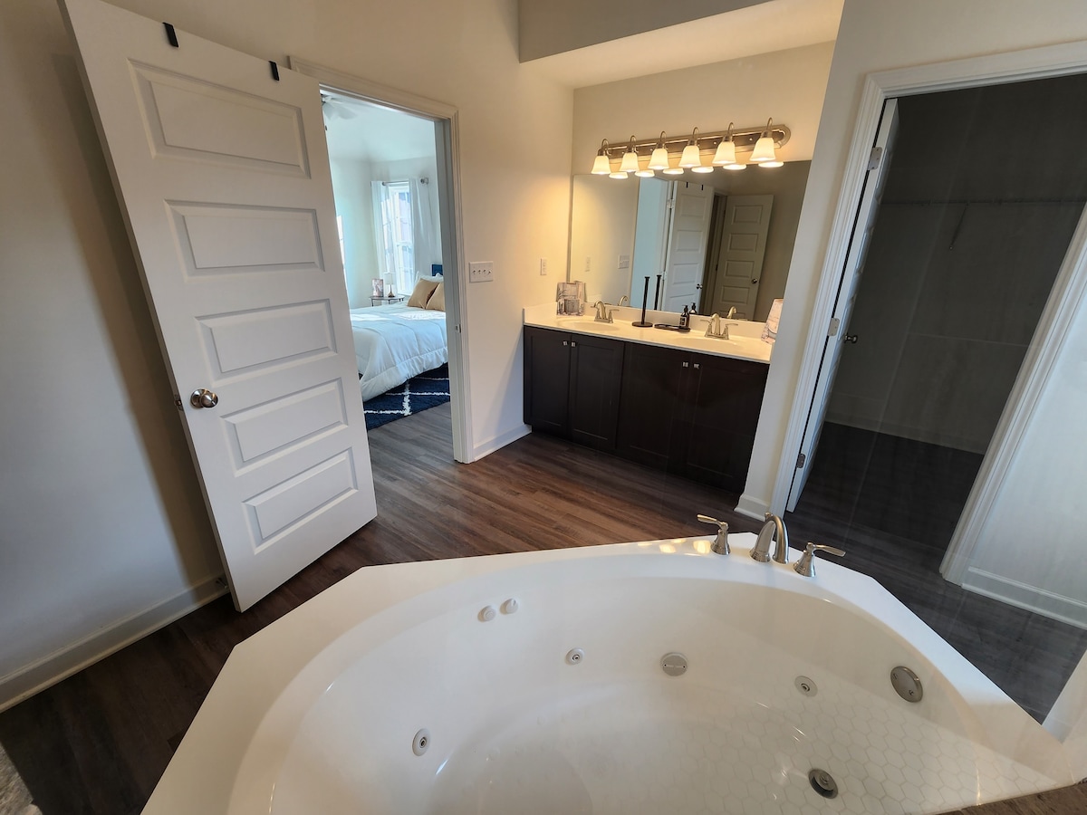 The spacious bathroom features a large jetted tub with chrome fixtures. Two sinks are positioned against a dark vanity. Natural light enters from an adjoining room, highlighting the modern flooring. A doorway leads to a secondary area, enhancing the open layout.