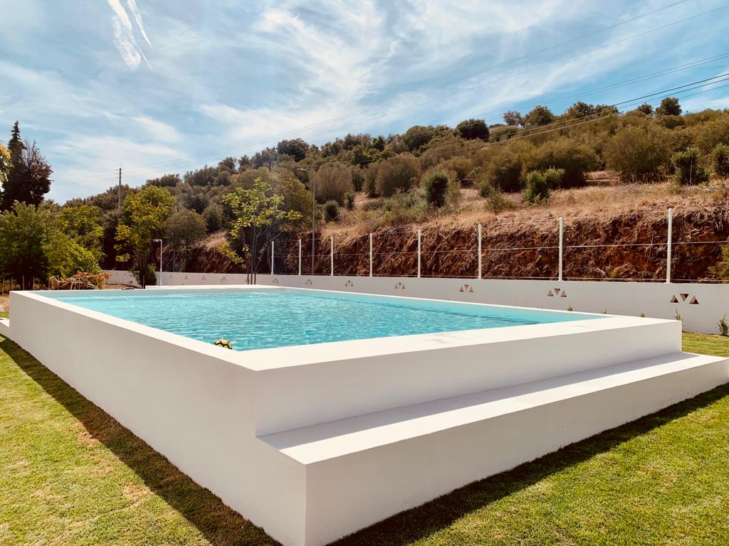 A refreshing outdoor swimming pool is framed by well-maintained greenery. The pool features steps leading into the water, with white walls surrounding the area. A gentle slope of hills can be seen in the background, enhancing the serene atmosphere.