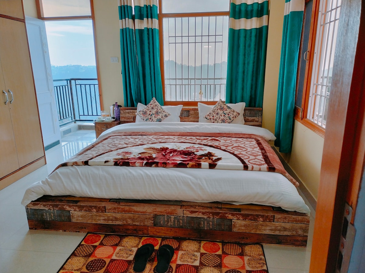 A spacious bedroom features a large bed with a patterned blanket and pillows adorned with floral designs. Floor-to-ceiling windows allow natural light to fill the room, showcasing panoramic views of the mountains. Vibrant curtains frame the windows, and a colorful rug adds warmth to the space.