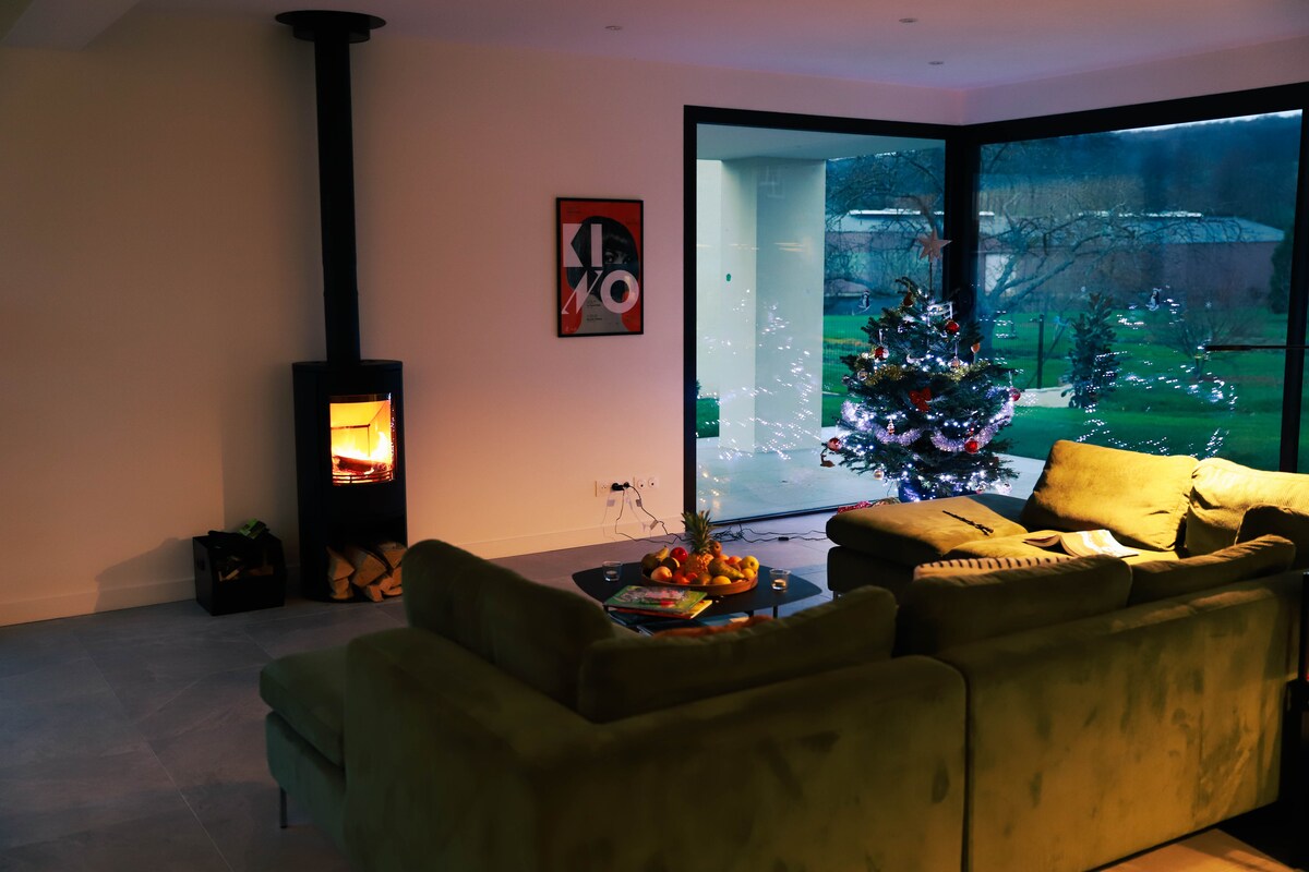 A spacious living area features a green sectional sofa arranged around a low table with a fruit bowl. A wood-burning stove adds warmth, and large glass doors provide a view of the garden and a decorated Christmas tree in the corner.