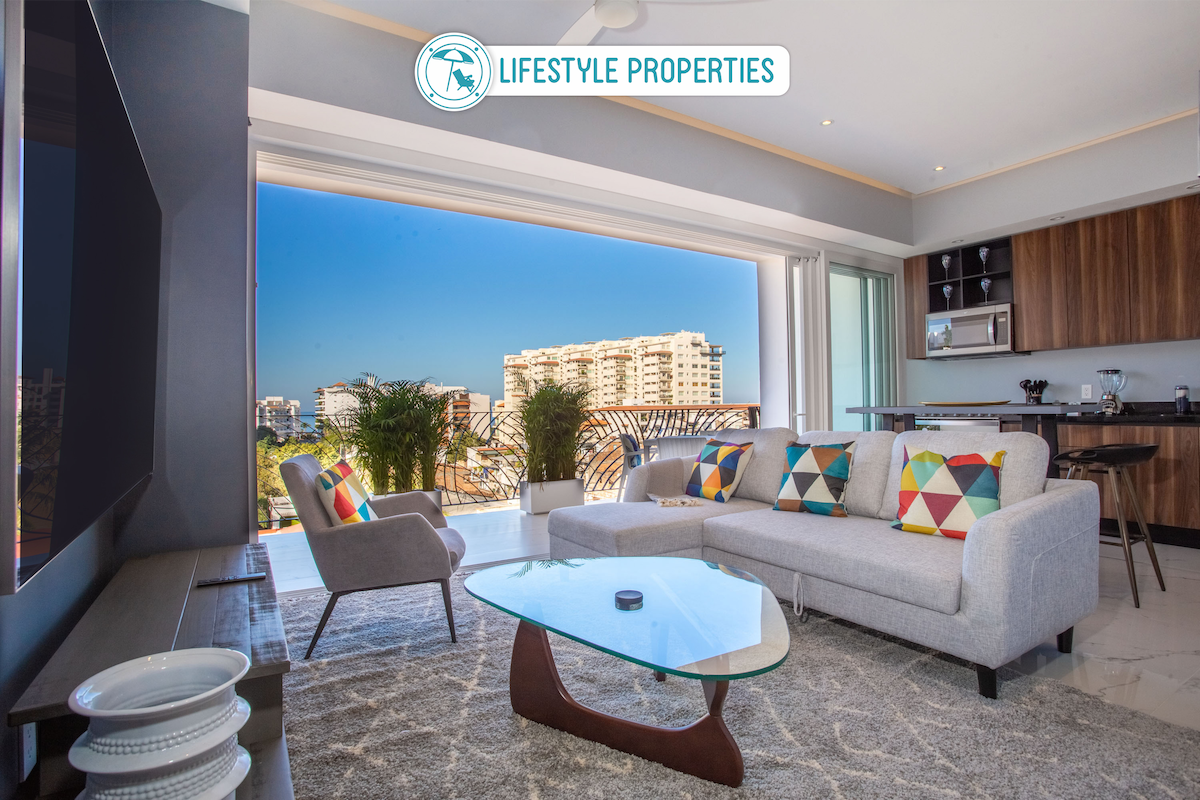 A modern living area features a comfortable grey sofa adorned with colorful cushions, paired with a glass coffee table. Expansive sliding doors provide access to a balcony with city views, while decorative plants add a touch of greenery to the space.