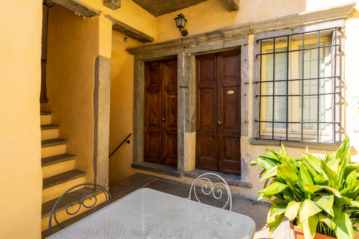 An inviting outdoor area features a metal table accompanied by decorative chairs, surrounded by vibrant greenery. To the left, a set of stairs leads to an upper level, while two wooden doors stand prominently under a warm yellow wall, enhancing the rustic charm.