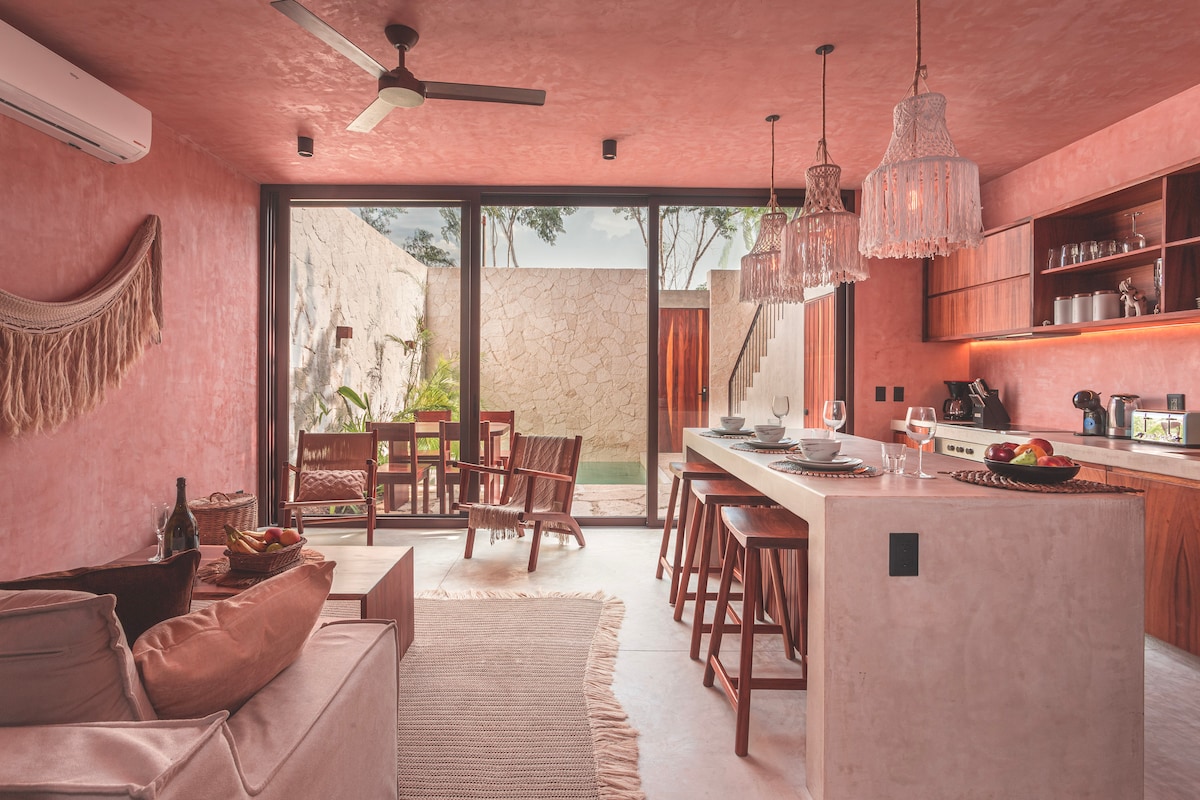 An open-concept living area features a cozy sofa, a dining space with wooden chairs, and a fully equipped kitchen with island seating. Natural light filters through expansive glass doors, leading to a private garden area, creating a seamless indoor-outdoor flow.