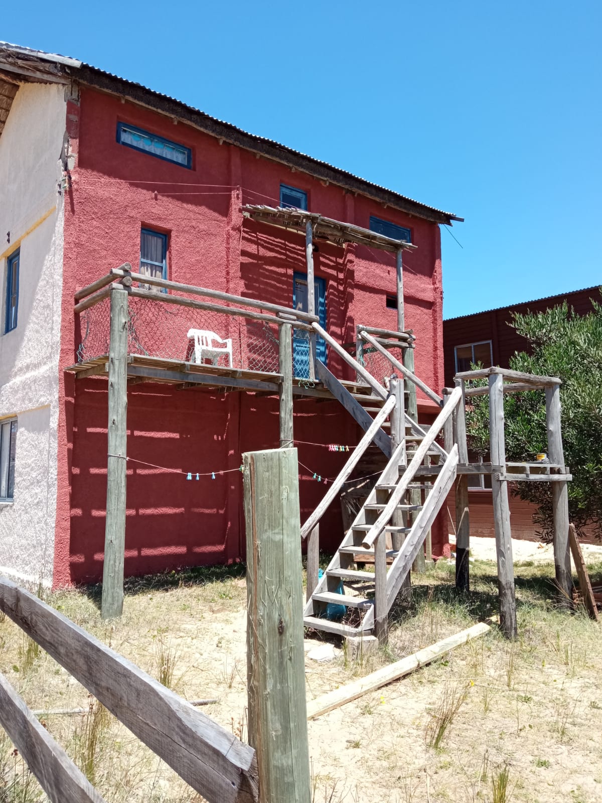 A two-story building features a red exterior with several large windows. A wooden staircase leads to a deck that provides elevated outdoor space, furnished with a white chair. Surrounding vegetation and rustic fencing add to the natural landscape.