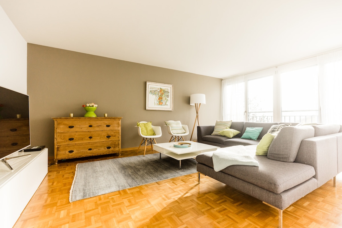 A bright and spacious living area features large windows allowing natural light to fill the room. A gray sectional sofa, accompanied by two accent chairs, is positioned around a central coffee table. A wooden sideboard and decorative artwork add visual interest to the space.