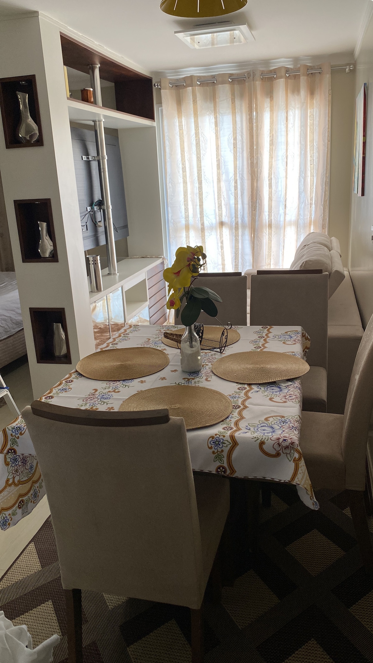 A dining area features a round table set with placemats and a floral centerpiece. Four plush chairs surround the table, complemented by a softly colored wall adorned with accent shelves. Natural light filters through sheer curtains, enhancing the cozy environment.