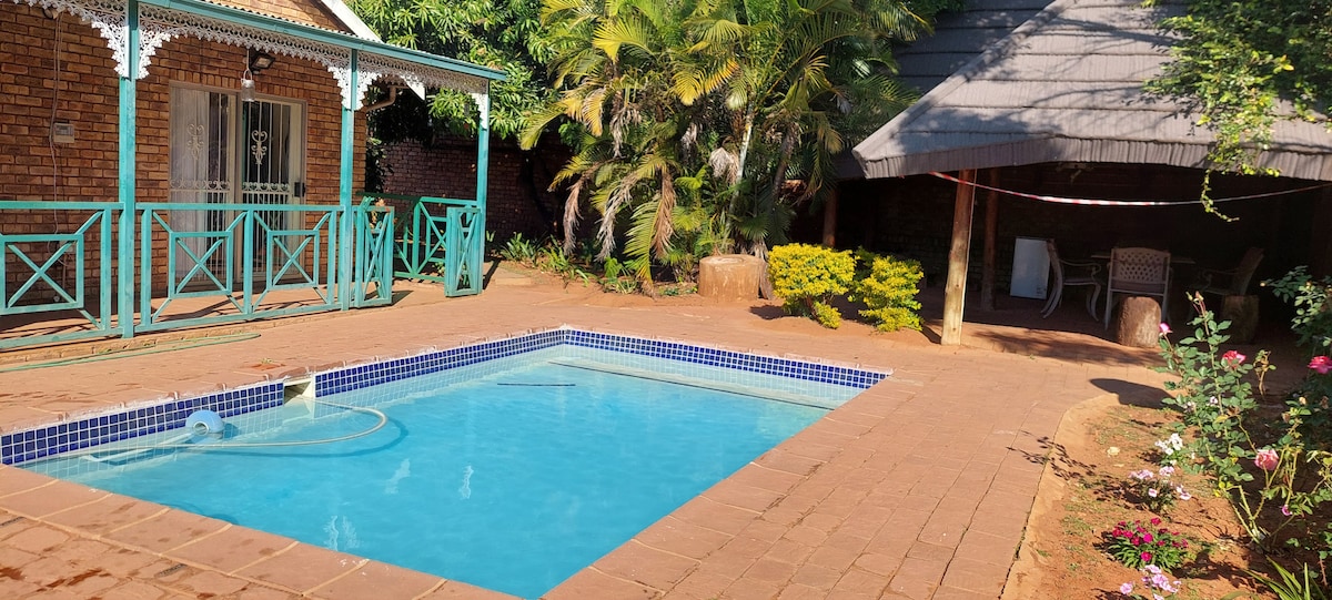 A backyard area features a refreshing swimming pool surrounded by a tiled patio. Lush greenery is visible, with a shaded seating area under a gazebo and decorative flowers lining the edge. Comfortable chairs are placed nearby, creating a relaxing outdoor environment.