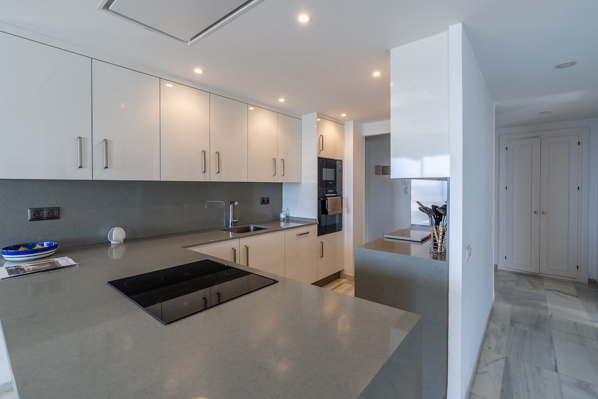 An elegant kitchen features sleek cabinetry with minimalistic handles and a spacious countertop. An induction cooktop is integrated into the surface, while modern appliances, including a built-in oven, are showcased. Neutral colors enhance the contemporary design, with natural light illuminating the area.
