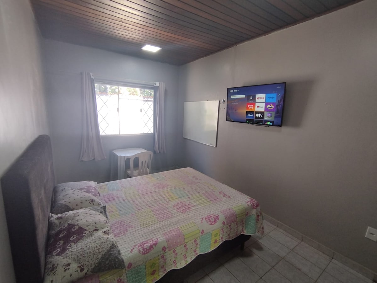 A cozy sleeping area features a spacious bed covered with a colorful patterned comforter. A modest white table and chair are positioned beside the bed. Natural light enters through the window adorned with light curtains, and a wall-mounted television is visible across the room.