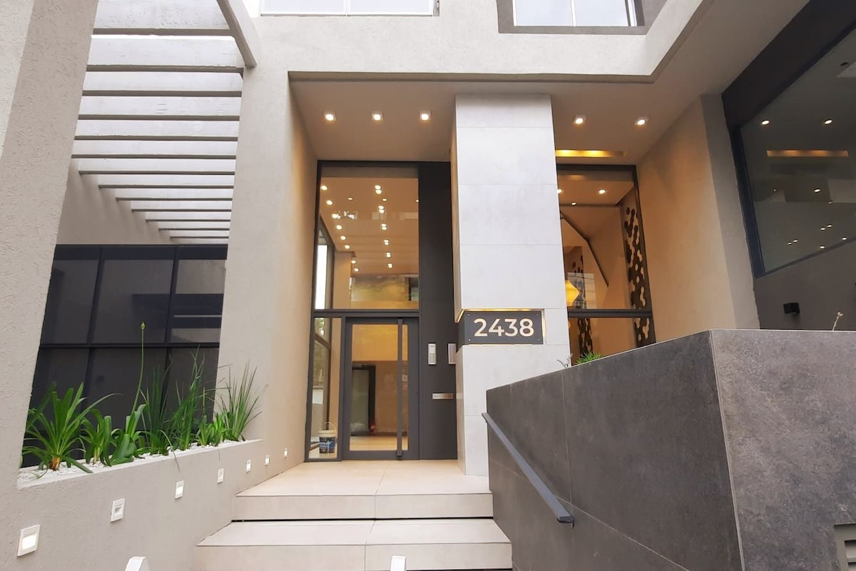 The building entrance features modern architecture with a sleek facade, highlighted by large glass doors and decorative elements. Soft lighting illuminates the pathway, while minimalist landscaping with green plants adds a touch of nature to the entryway.