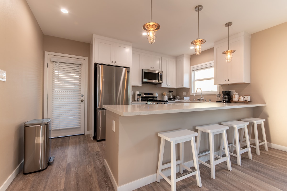 The kitchen features modern cabinetry and stainless steel appliances. A large island with seating for four is present, illuminated by hanging light fixtures. A door leads to an outdoor area, and a silver trash can complements the space.