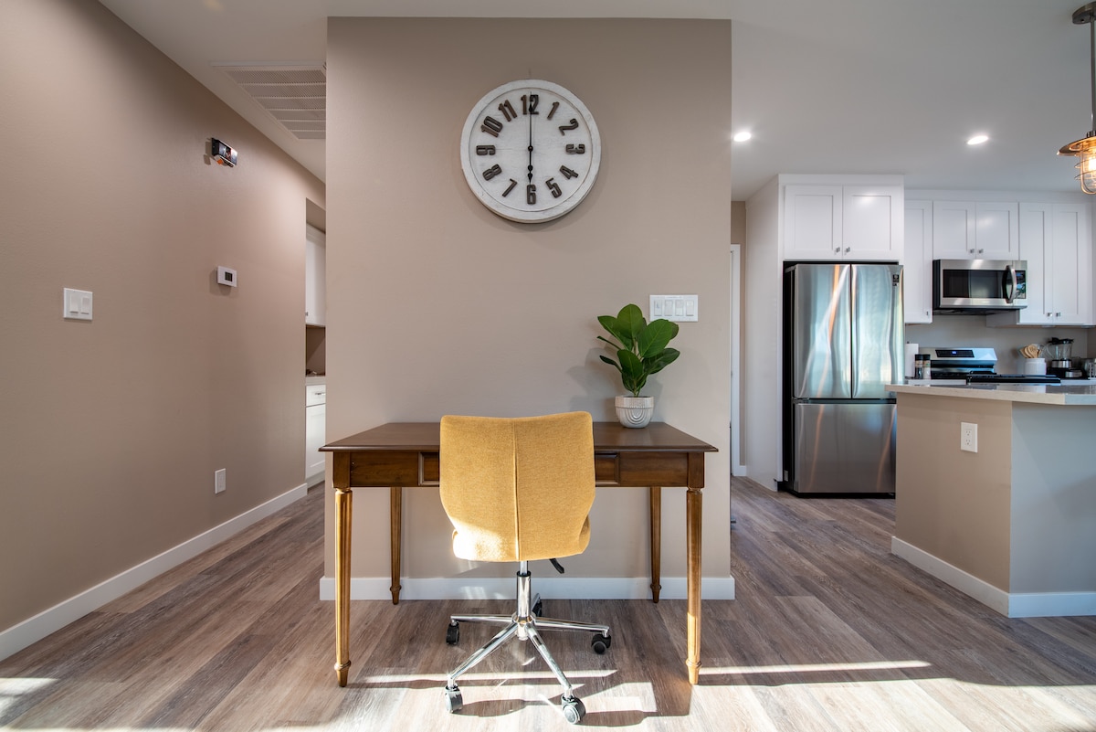 A functional workspace is positioned against a neutral wall, featuring a wooden desk with a yellow chair. A round clock is mounted above the desk. In the background, a modern kitchen with white cabinetry and stainless steel appliances is visible, enhancing the overall brightness of the space.