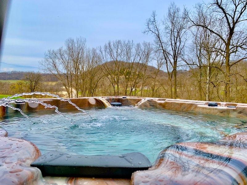An outdoor hot tub is positioned with a view of rolling hills and trees. Gentle water jets create soft splashes in the tranquil water, enhancing the serene natural setting.