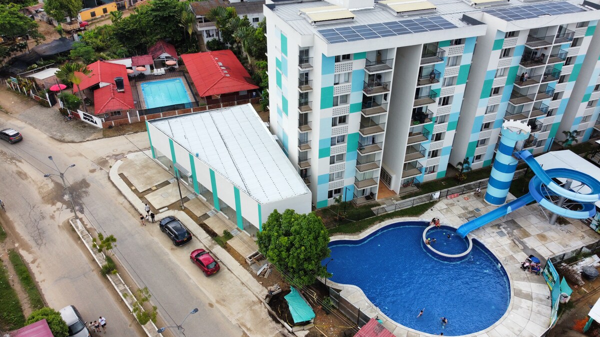 An aerial view captures a modern multi-story building alongside a vibrant blue swimming pool featuring a water slide. Nearby, various structures are visible, including red-roofed buildings and parking spaces, offering a sense of the surrounding community.