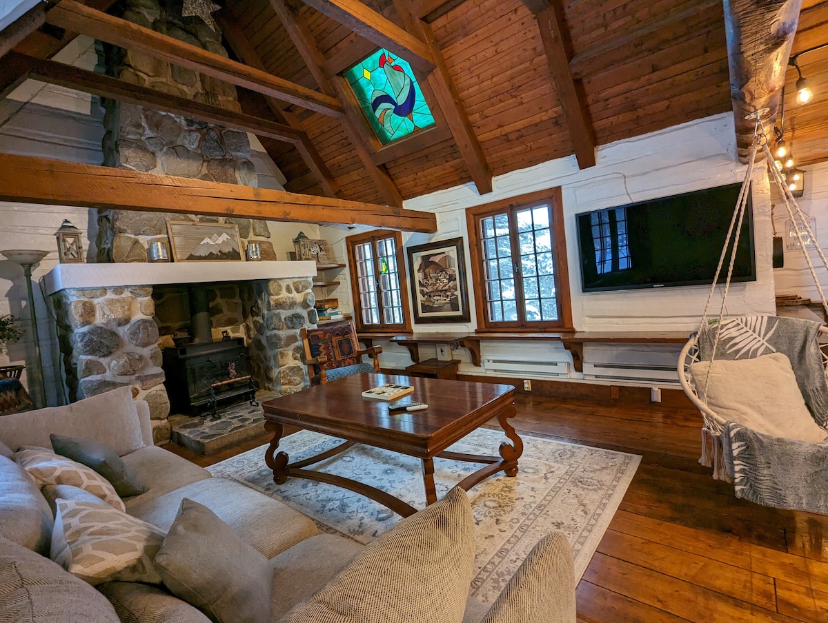 A living room is showcased with a large stone fireplace as the focal point, complemented by exposed wooden beams and a cathedral ceiling. A comfortable sofa surrounds a wooden coffee table, while light streams through multiple windows adorned with decorative treatments.