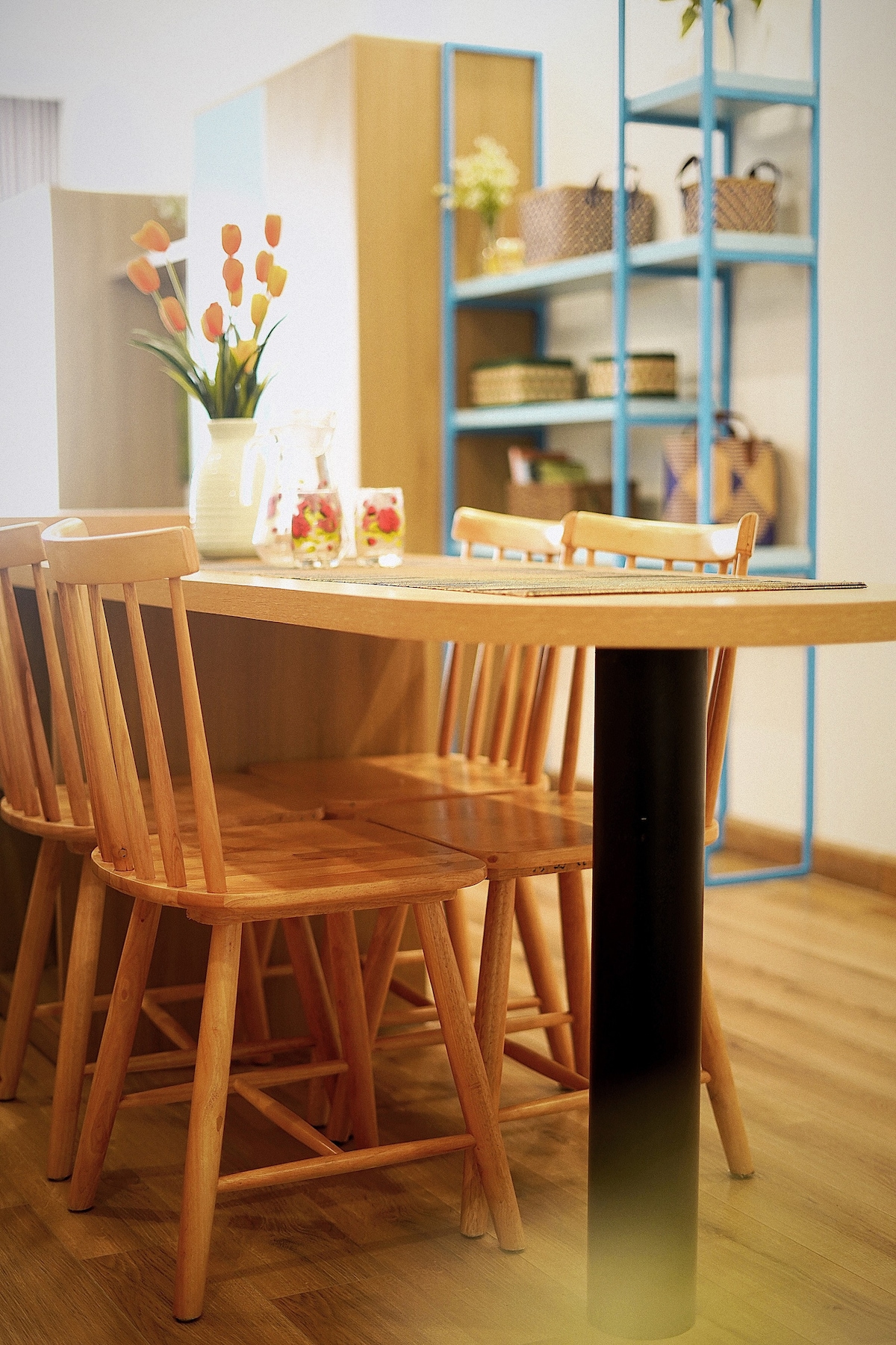 A dining area is presented with a wooden table and several light-toned wooden chairs. Bright orange tulips are positioned in a vase on the table, while a shelf in the background displays various baskets and decor items.