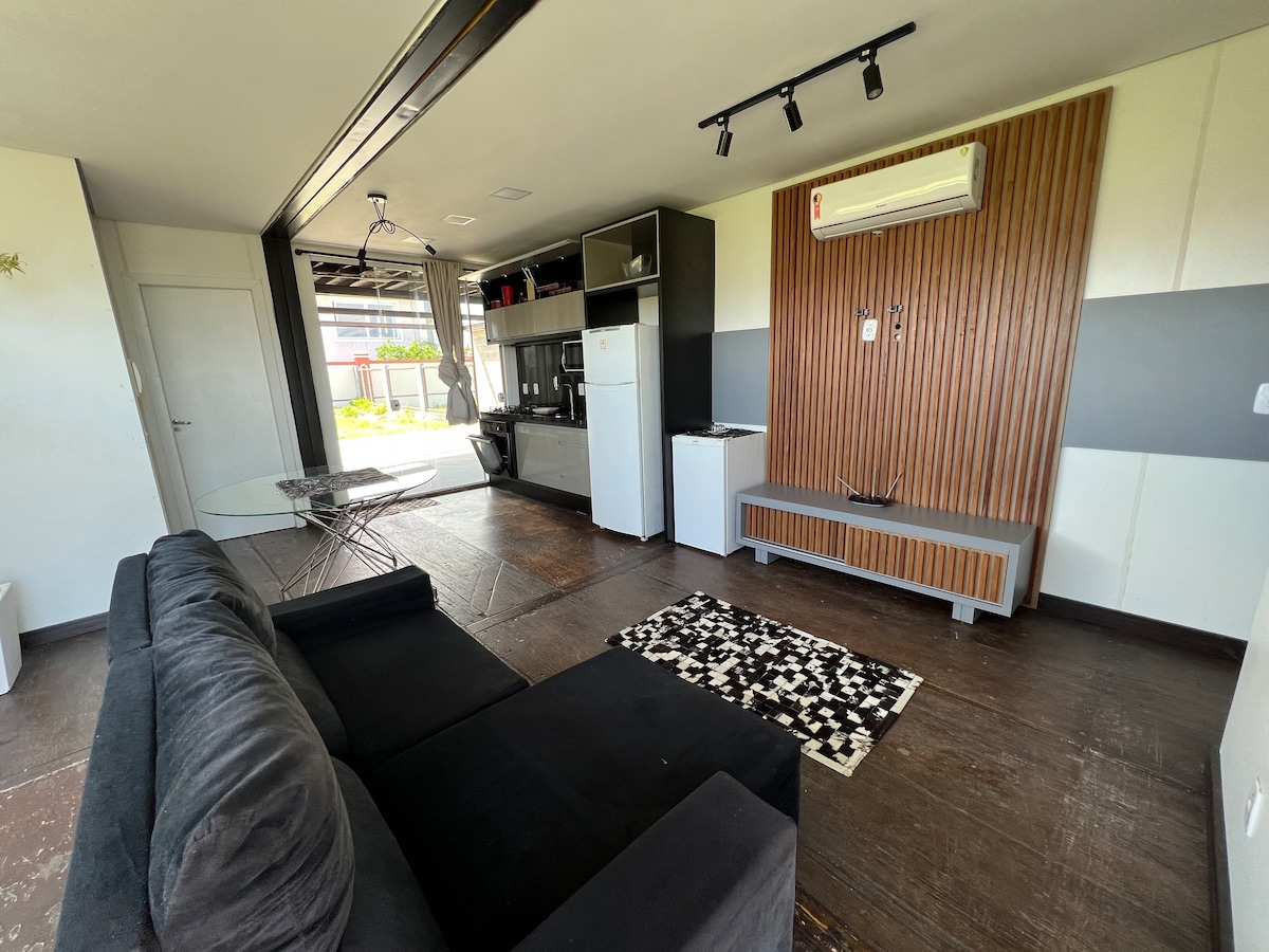 An open living area is presented with a dark sofa facing a wooden accent wall. A modern kitchenette and dining space are visible to the side. Large sliding doors provide access to the outdoor area, with sunlight illuminating the space.