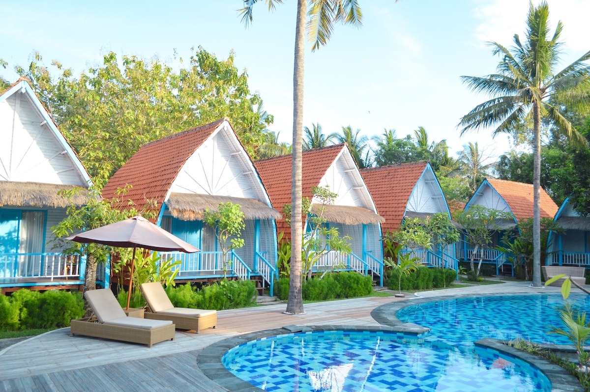 A collection of bungalows with thatched roofs are surrounded by lush greenery and palm trees. An inviting outdoor swimming pool with a distinct blue pattern reflects the clear sky, while comfortable lounge chairs are positioned nearby for relaxation.
