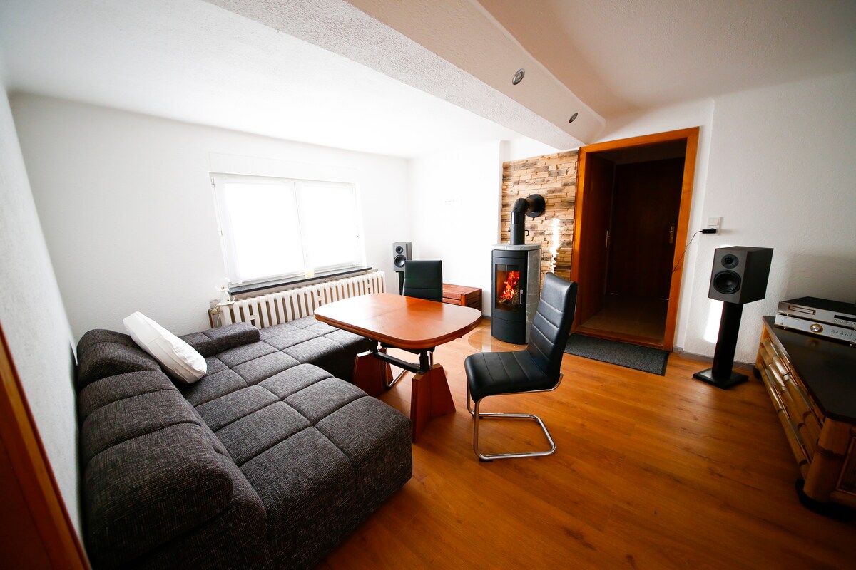 A modern living area features a sectional sofa in a neutral tone, paired with a sleek dining table. A contemporary chair complements the space. Large windows allow natural light to fill the room, highlighting the warm wood flooring and unique fireplace design.