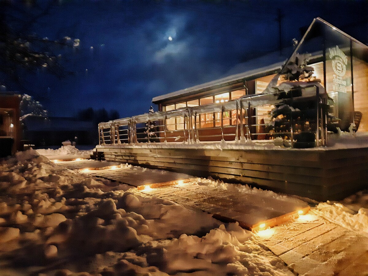 A well-lit terrace is displayed, surrounded by a blanket of snow. The structure features large windows that reflect the evening light, creating a cozy ambiance amidst a serene winter landscape. Softly glowing lights line the snowy path, inviting guests to explore.