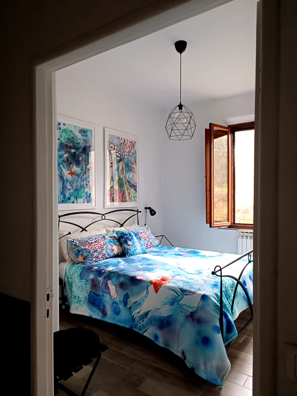 A serene bedroom showcases a metal bed adorned with a colorful bedspread featuring blue and white patterns. Two framed artworks decorate the walls, and natural light filters through a window with a wooden frame, creating a tranquil ambiance.