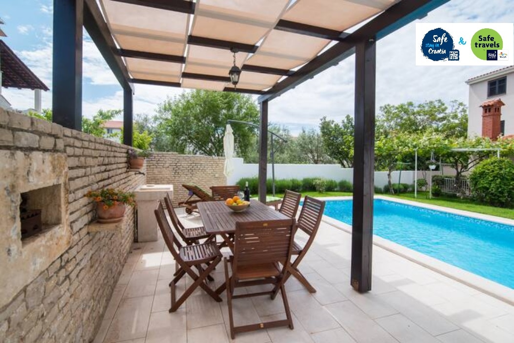 An outdoor seating area is shown beneath a wooden pergola, featuring a rectangular dining table surrounded by six folding chairs. A stone barbecue is visible nearby, and a refreshing pool is located just beyond, framed by lush greenery and well-maintained landscaping.